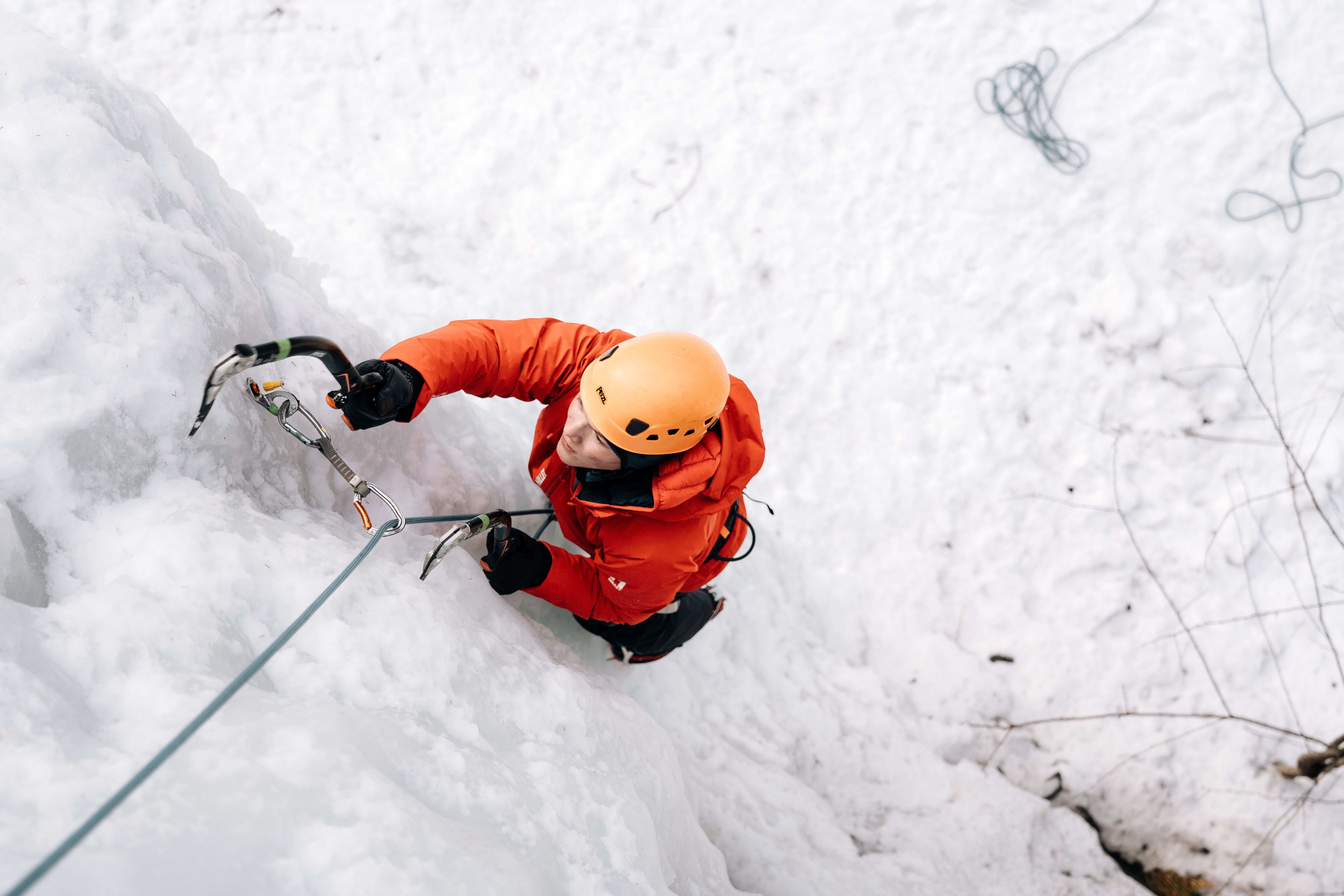 Person climbing a frozen waterfall with a helmet and ice pick; safety rope visible.